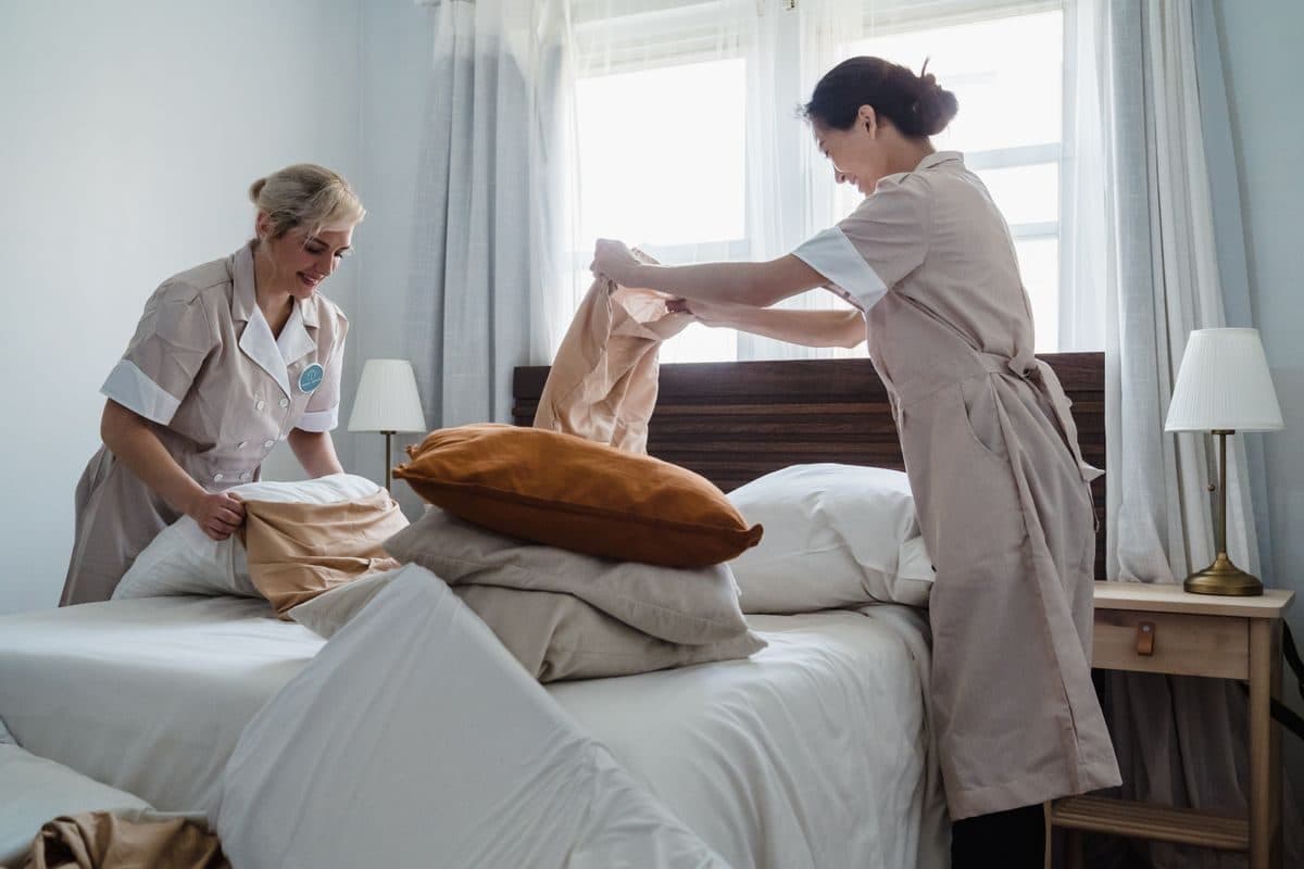 Two uniformed housekeepers making up a bed in a sunlit hotel room