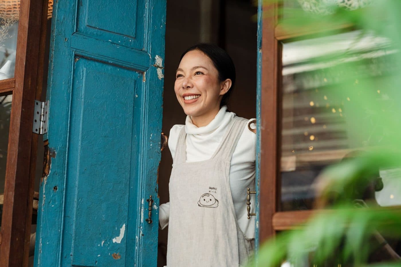 A shop owner in an apron smiling at the front door of her store